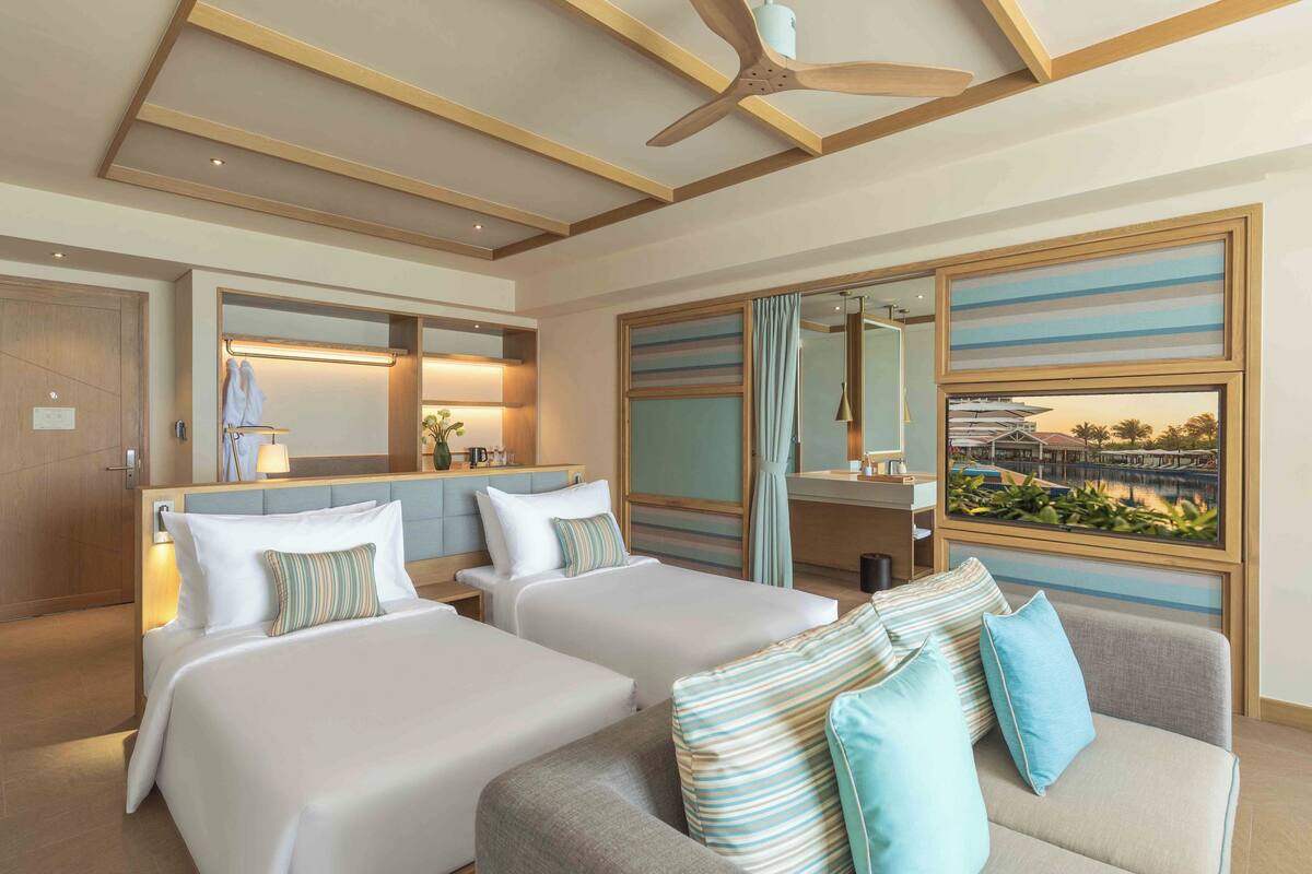 A hotel room with two beds, a sofa, a ceiling fan, a large window with a view, and a bathroom with a mirror and towel rack.