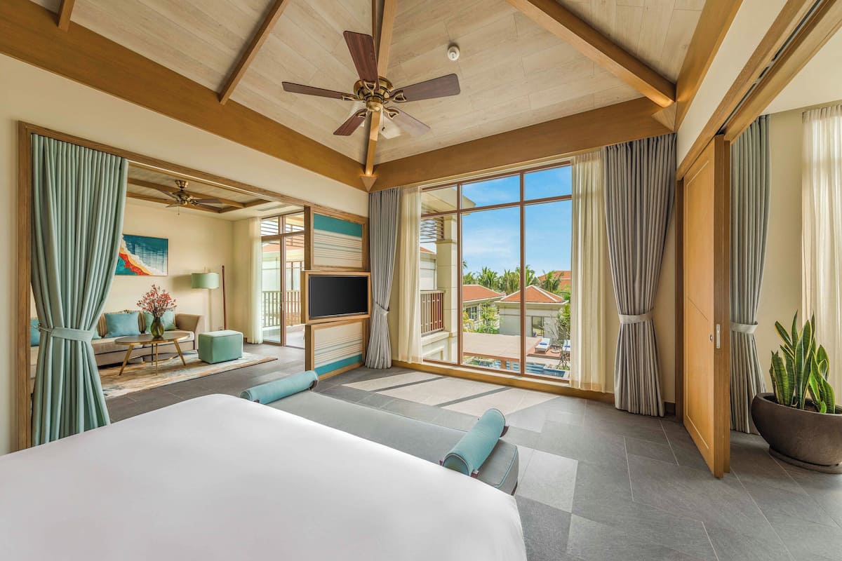 A spacious hotel room with a large bed, a ceiling fan, a view of the outdoors, and a balcony with curtains.