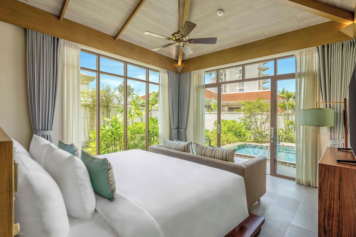 A hotel room with a large bed, a ceiling fan, and a view of a swimming pool and greenery.