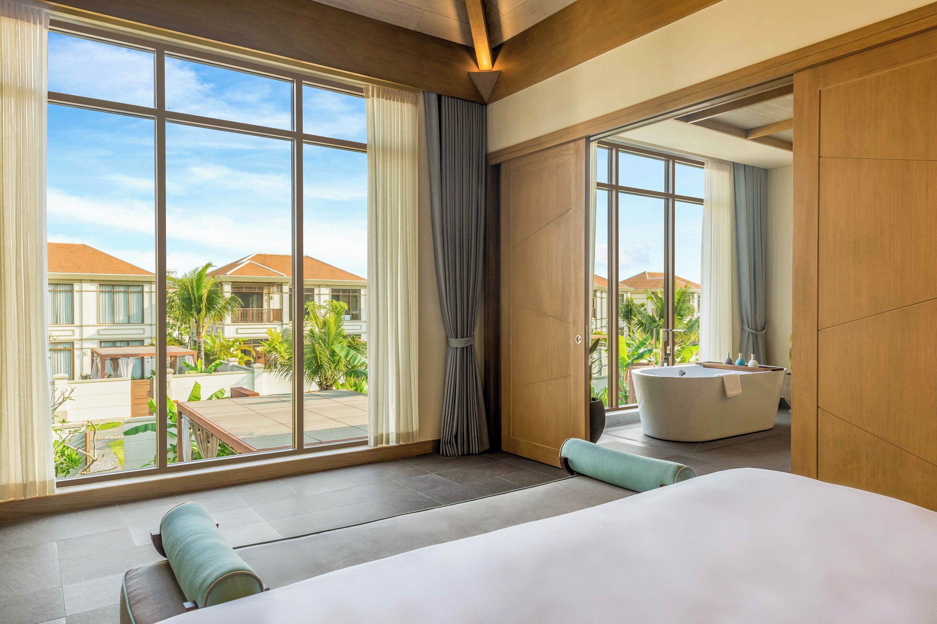 Ein modernes Hotelzimmer mit großen Fenstern, einer Badewanne und Blick auf die Gebäude und Grünflächen des Resorts.