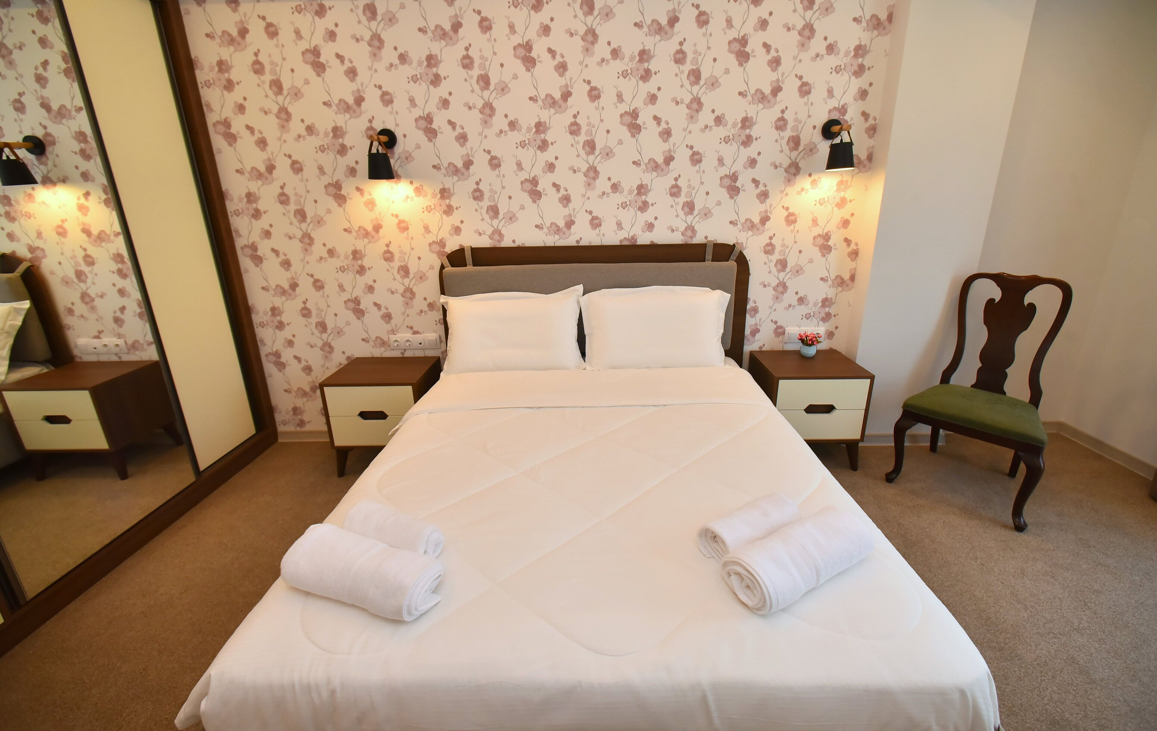 A neatly made bed with two pillows and two rolled towels, a wooden nightstand with a lamp, a chair, and a mirror on the wall.