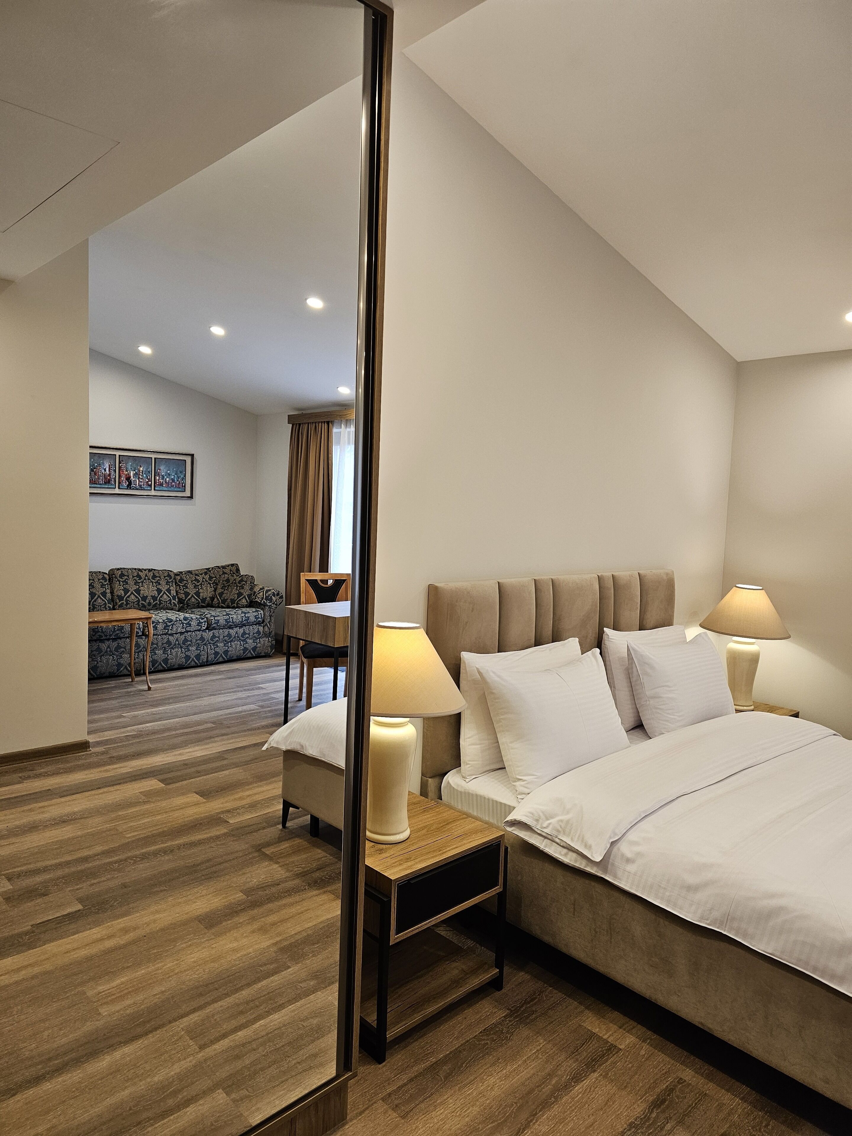 Habitación de hotel con una cama grande, dos lámparas de noche, una mesita de noche de madera, un sofá y una mesa de comedor al fondo.