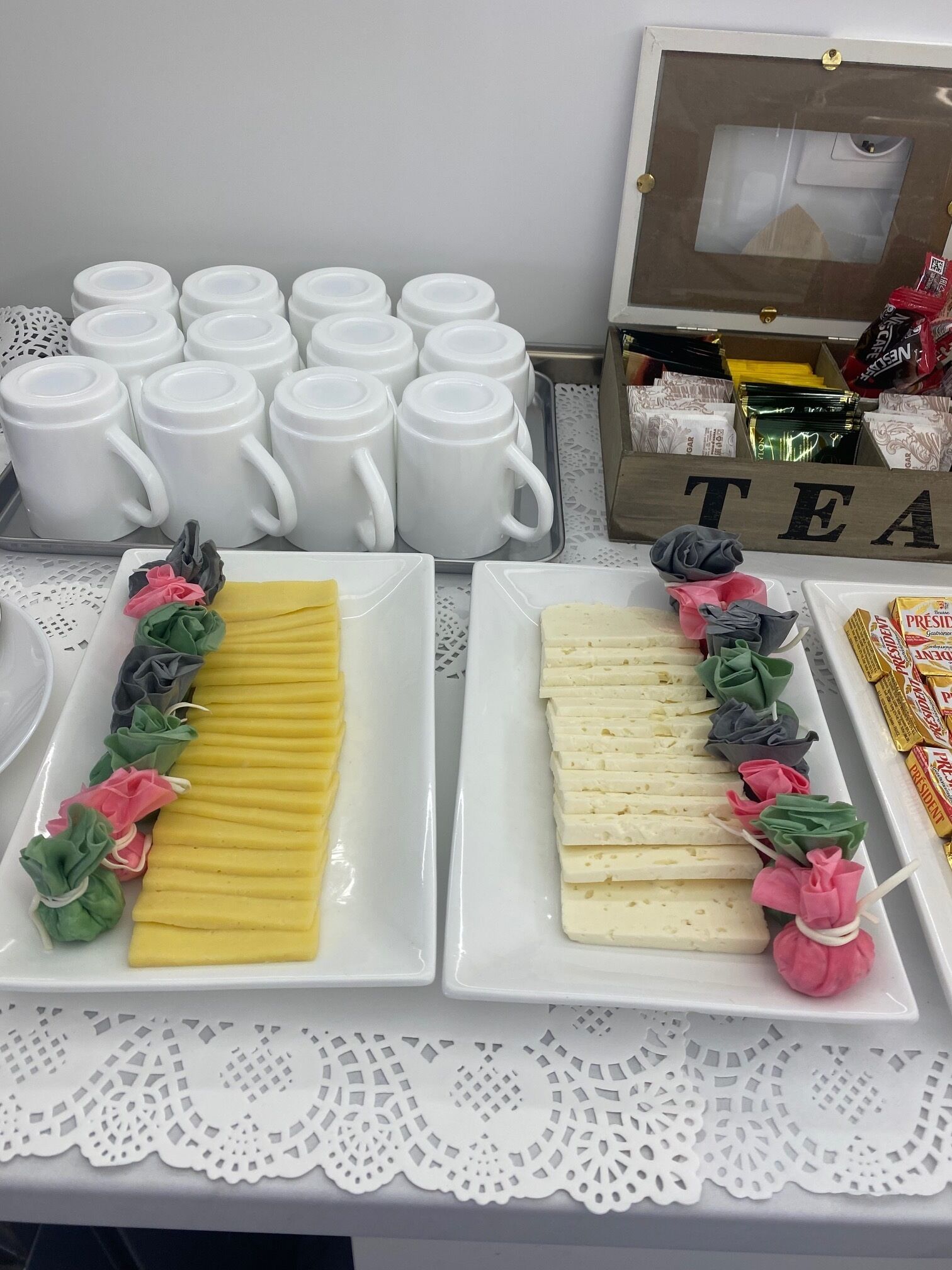 Una mesa de buffet con tazas blancas, una bandeja de queso amarillo y blanco, y una bandeja de queso blanco y rosa.