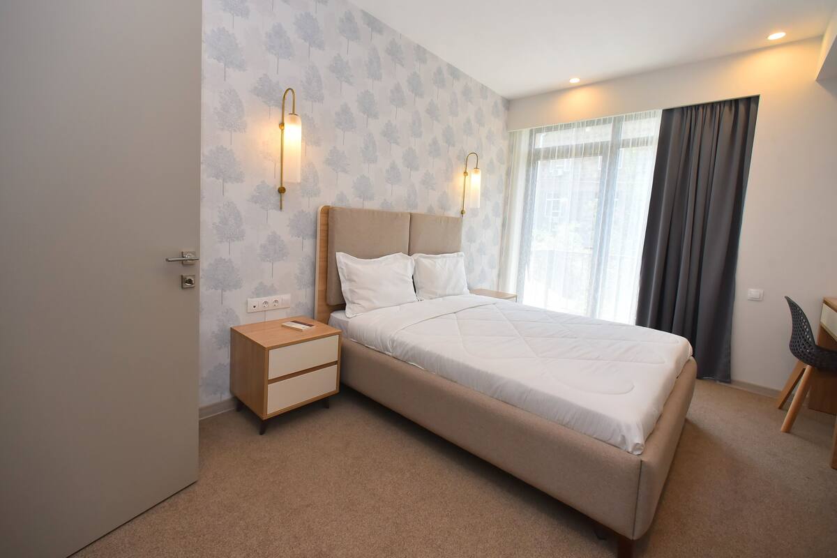 A modern hotel room with a large bed, a bedside table, a chair, and a window with curtains.