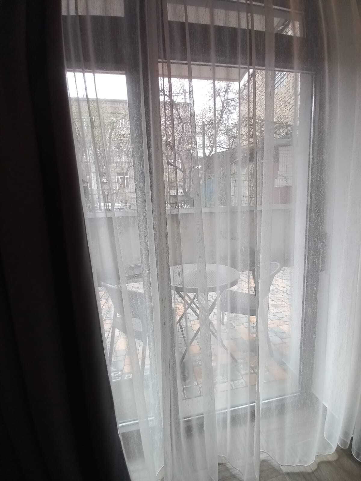 A room with a large window covered by sheer curtains, a small round table, and a chair visible through the window.