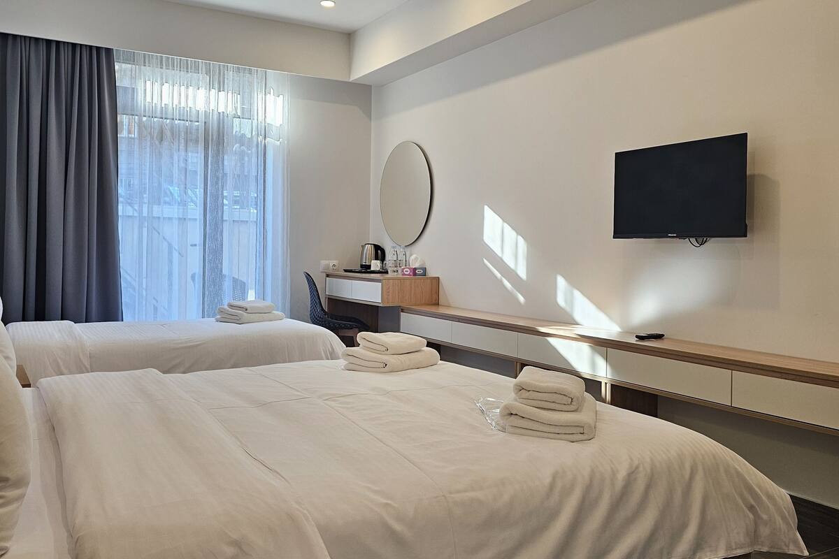 A modern hotel room with two beds, a desk, a flat-screen TV, and a large window with curtains.