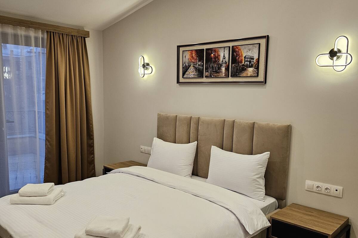 A neatly made bed with white linens and two pillows, a bedside table with a lamp, and a framed picture above the bed.