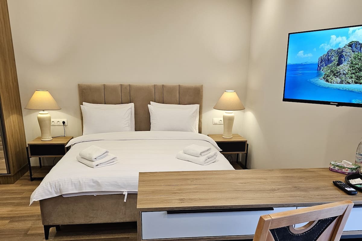 A hotel room with a bed, two bedside lamps, a flat-screen TV, a wooden desk, and a chair.
