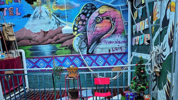 Une fresque colorée représentant un toucan et un paysage de montagne, une table à manger avec des assiettes remplies de nourriture et des boissons, et un petit sapin de Noël.