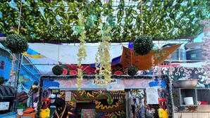 Un étal de marché en plein air, aux couleurs vives, agrémenté de plantes suspendues, d’un motif décoratif représentant un soleil et proposant divers articles à la vente.