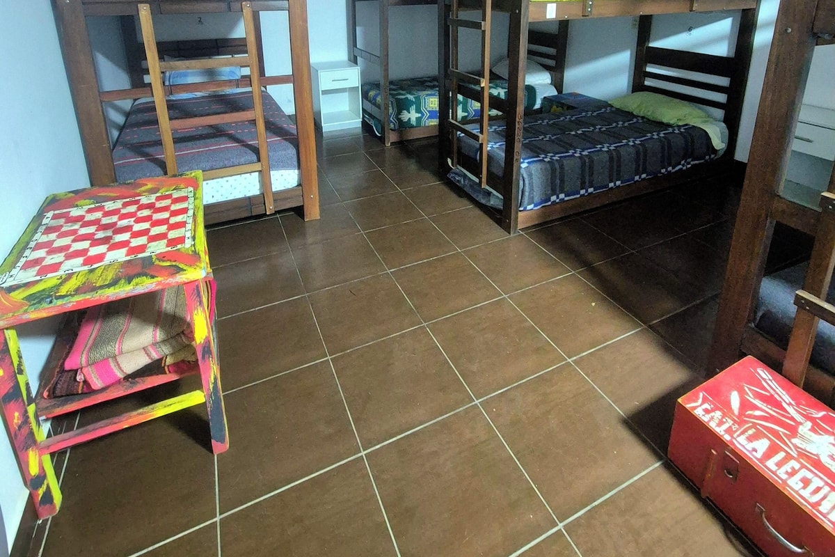 A dormitory room with bunk beds, a small table with a checkered tablecloth, and a red suitcase.