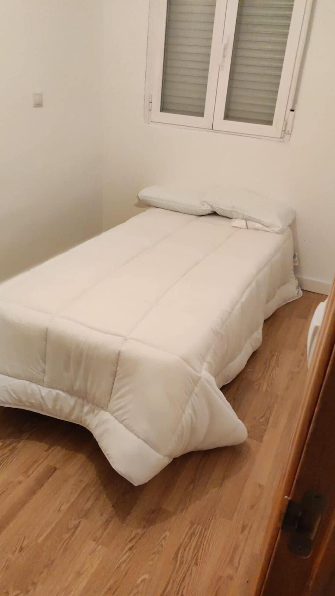 A small, single-bed room with a wooden floor, white walls, and a window with blinds.