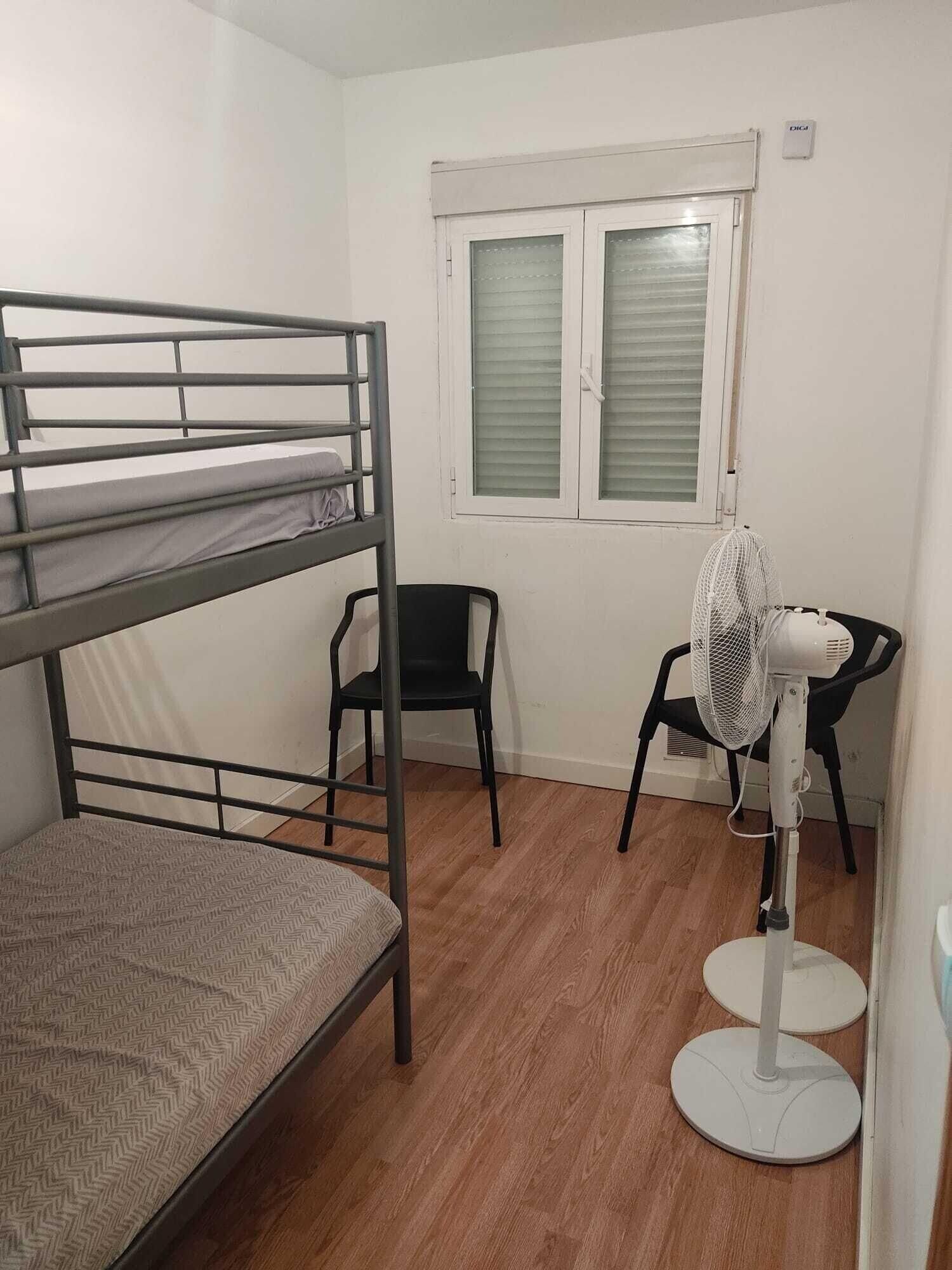 Una cama individual superpuesta, un escritorio con silla, un ventilador de pie y una ventana con persianas.