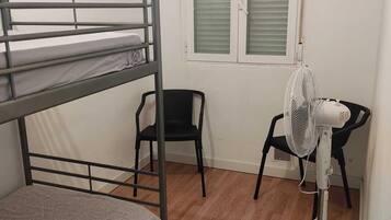 Una cama individual superpuesta, un escritorio con silla, un ventilador de pie y una ventana con persianas.
