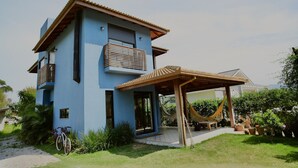 A modern two-story house with a blue exterior, a wooden balcony, and a covered patio area with a hammock. - Refúgio da Ibiraquera Jacuzzi e Vista (Imbituba)