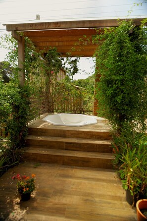 Una pérgola de madera con un jacuzzi debajo, rodeada de exuberante follaje y escalones que conducen al jacuzzi.