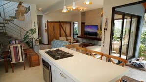 A modern kitchen with a central island, wooden dining table, and chairs. There is a TV mounted on the wall and a staircase leading to another level. - Refúgio da Ibiraquera Jacuzzi e Vista (Imbituba)