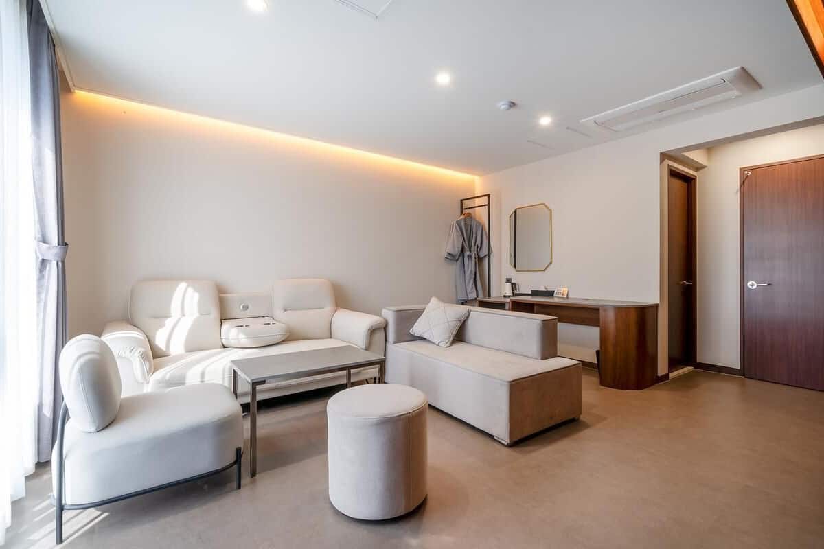 A modern hotel room with a sofa set, a coffee table, a small ottoman, a vanity with a mirror, and a wooden door.