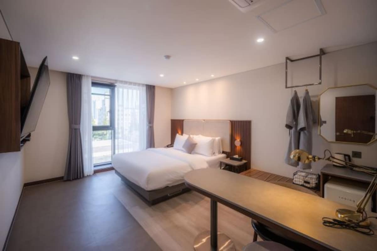 A hotel room with a large bed, a desk, a chair, a desk lamp, a mirror, and a window with curtains.