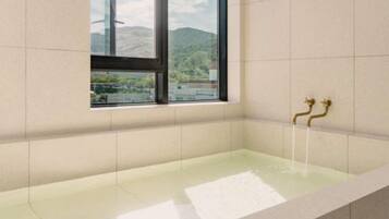 A bathroom with a bathtub, a window with a view of greenery and mountains, and a gold faucet.