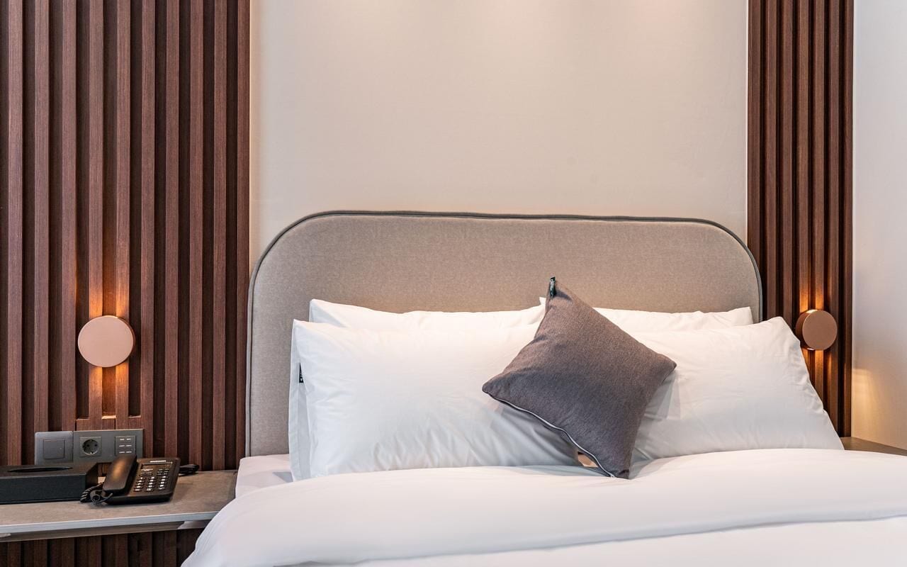 A modern hotel room with a bed, a bedside table with a phone, and two wall-mounted lamps.