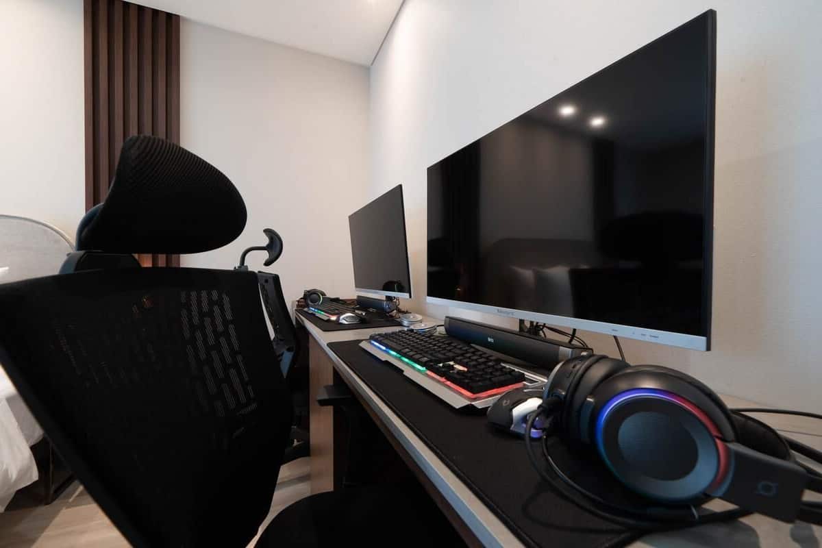A hotel room with a bed, a desk with a computer setup including a monitor, keyboard, and headphones, and a large window with blinds.
