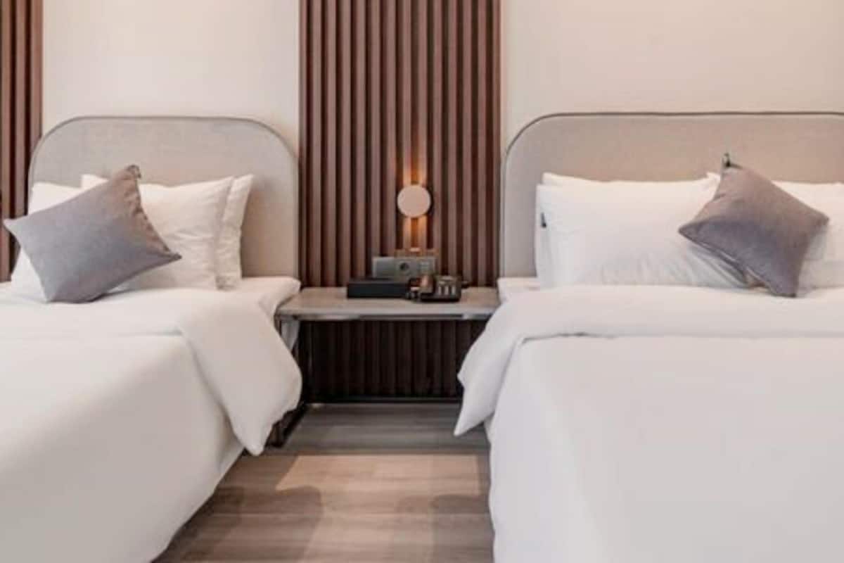 A hotel room with two beds, each with white bedding and grey pillows. There is a small table with a lamp and a radio between the beds.