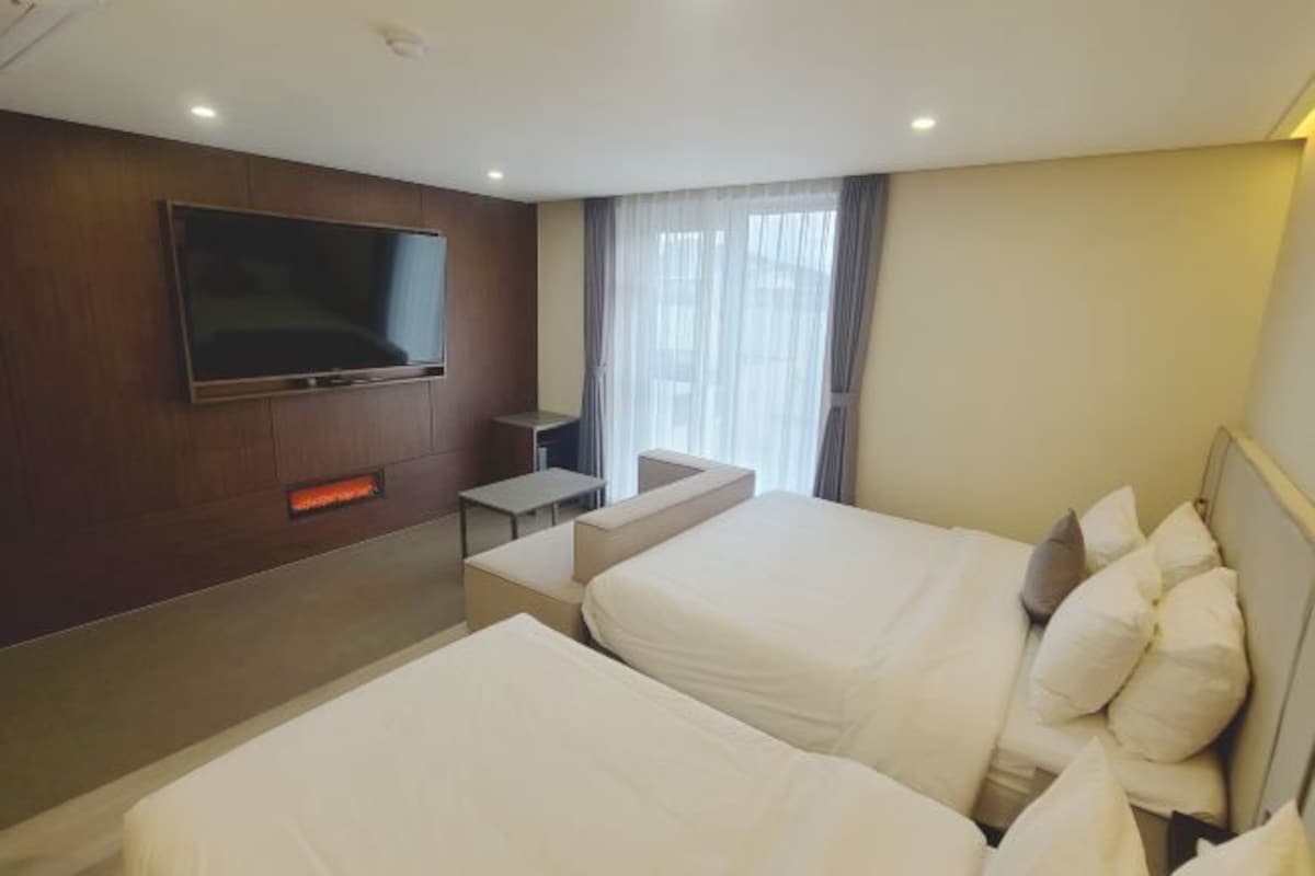 A hotel room with two beds, a TV mounted on the wall, a small table, and a window with curtains.