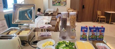 Montaje de un bufé de desayuno con diversos alimentos y un área de personal al fondo.