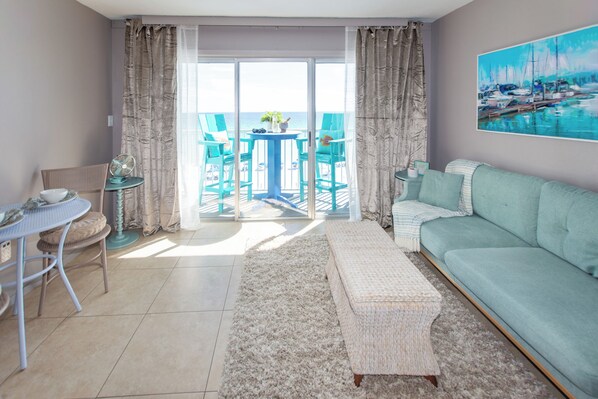A living room with a sofa, a table, chairs, and a view to the outside. - 🌊Islander 502 - The Sea Glass Studio | Gulf Views | Pool🌊 (Fort Walton Beach)