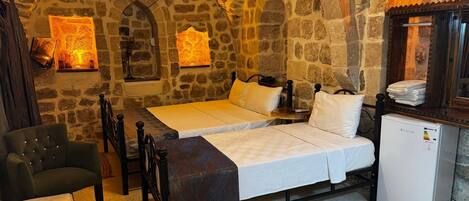 A stone-walled room with two beds, a chair, a small table, and a refrigerator.