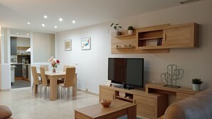 Un salon moderne avec un téléviseur, des meubles en bois, un coin repas et une cuisine en arrière-plan.