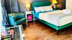 A hotel room with a green upholstered chair, a bed with a white and patterned bedspread, a nightstand with a lamp, and a framed floral artwork on the wall. - New Luxury apt,Javits Center,NYC (New York)