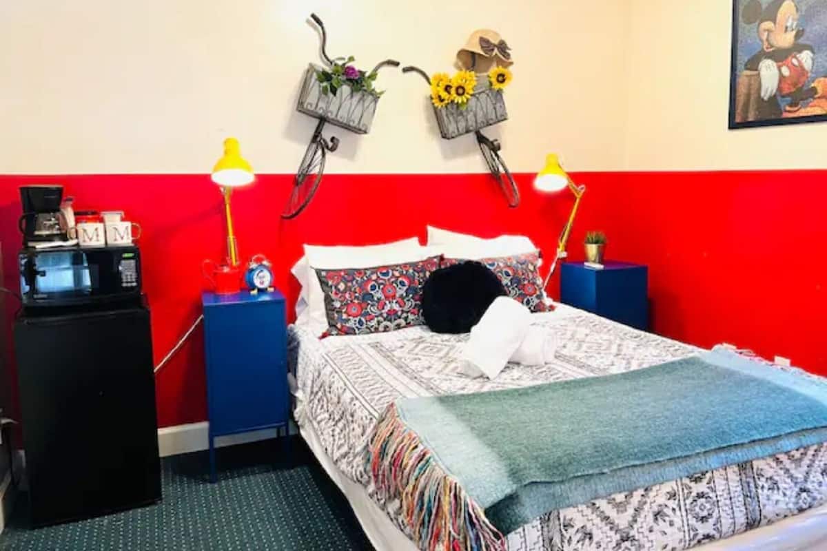 Ett sovrum med en färgskala i rött och vitt, en säng med mönstrat sänglinne, två väggmonterade lampor, en klocka och ett litet bord med en lampa och en växt.