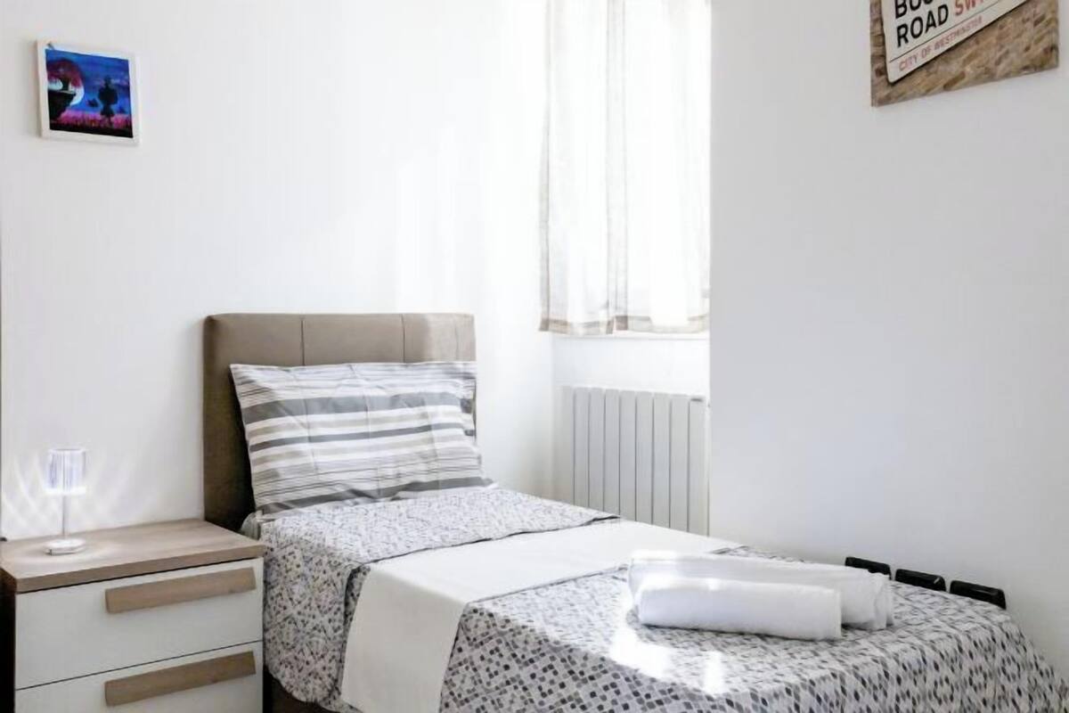 A bedroom with a bed, a bedside table, a lamp, a window with curtains, and a wall sign.