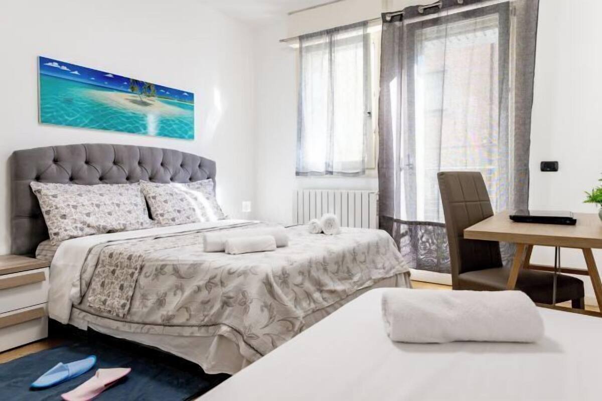 A bedroom with a bed, a desk, a chair, a window with curtains, and a painting on the wall.