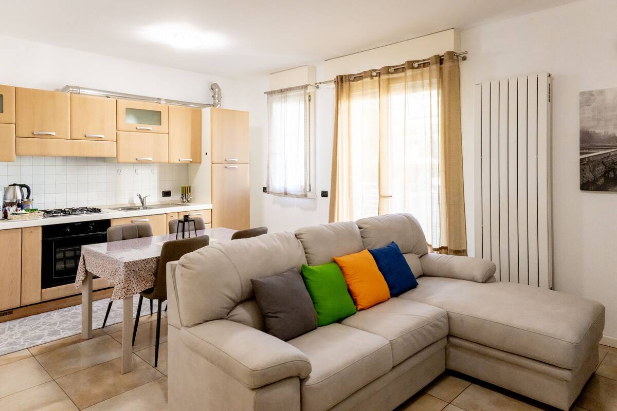A modern kitchen with wooden cabinets, a dining table, and a beige sofa with colorful cushions.