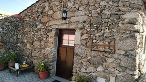 Edifício de pedra com porta de madeira, plantas em vasos e uma pequena área externa com mesas e cadeiras.