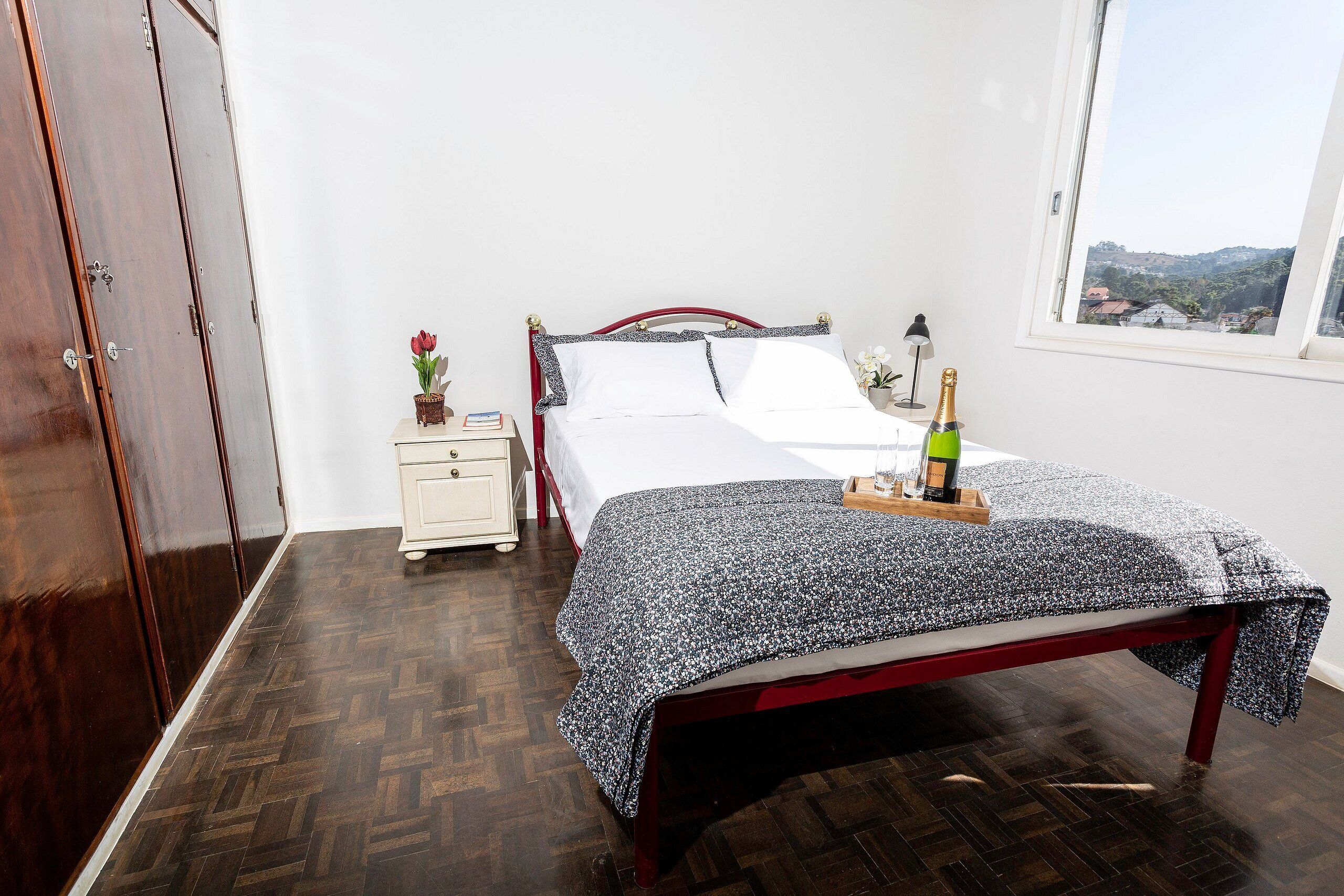 Quarto com piso de madeira, cama com colcha florida, mesa de cabeceira com um vaso de flores e janela com vista para as colinas.
