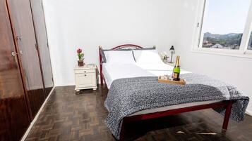 Quarto com piso de madeira, cama com colcha florida, mesa de cabeceira com um vaso de flores e janela com vista para as colinas.