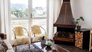 A cozy living room with a fireplace, a white furry chair, a coffee table with a plant, and a view through a large window. - Incrível Vista com 4 Quartos e Lareira (Campos do Jordão)