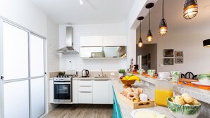 A modern kitchen with white cabinets, a built-in oven, a sink, and a countertop with various food items and a fruit bowl. - Incrível Vista com 4 Quartos e Lareira (Campos do Jordão)