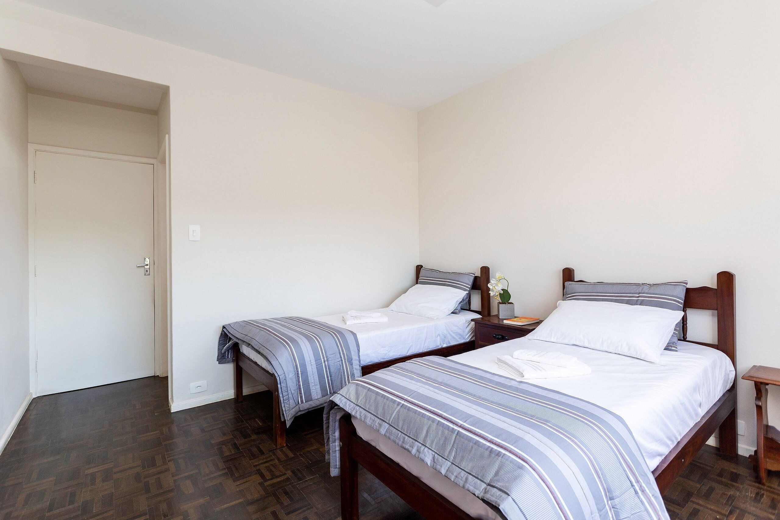 A room with two beds, a wooden floor, a white door, and a small table with a lamp.