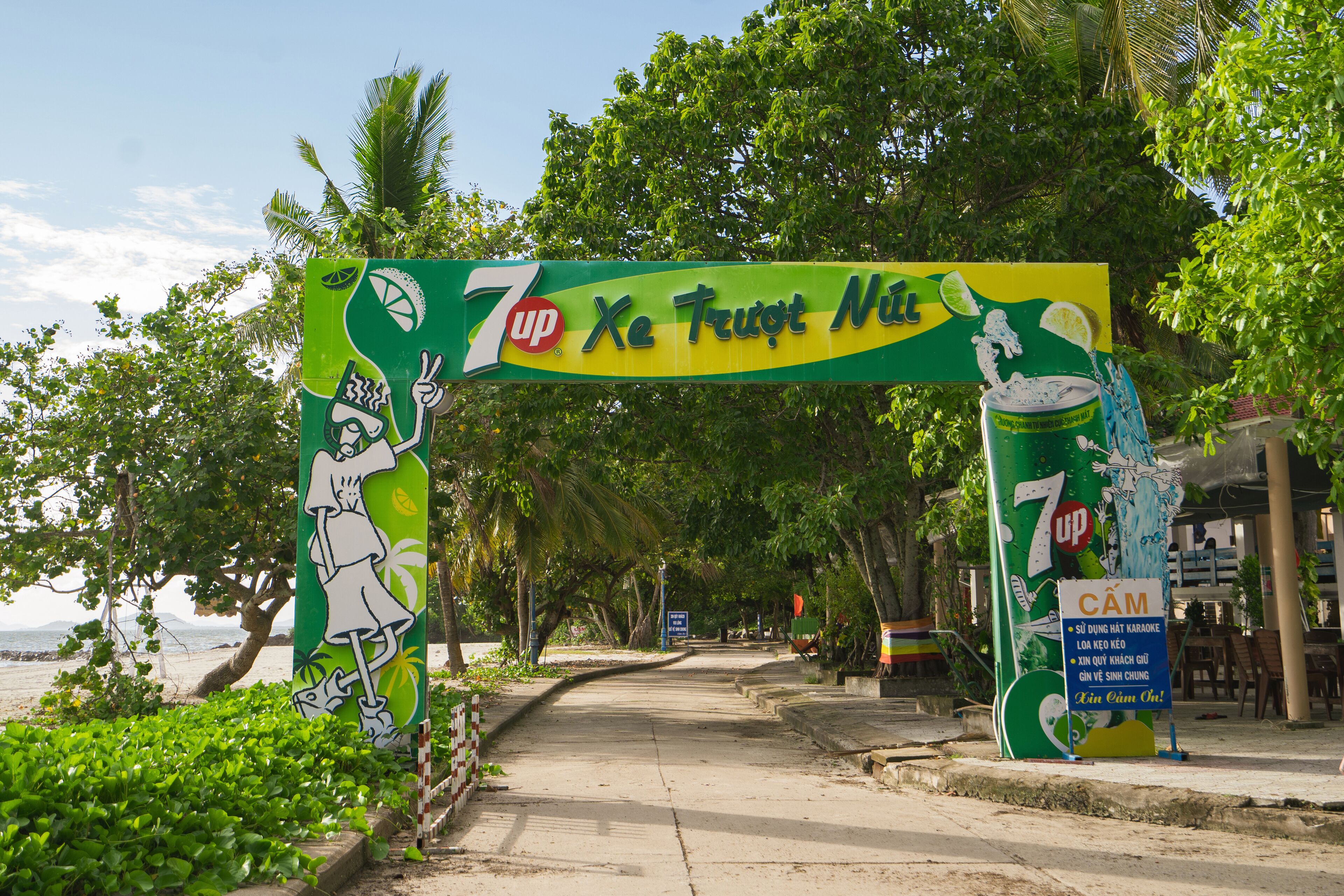 A 7UP branded archway at a beachside location with palm trees and a view of the sea.