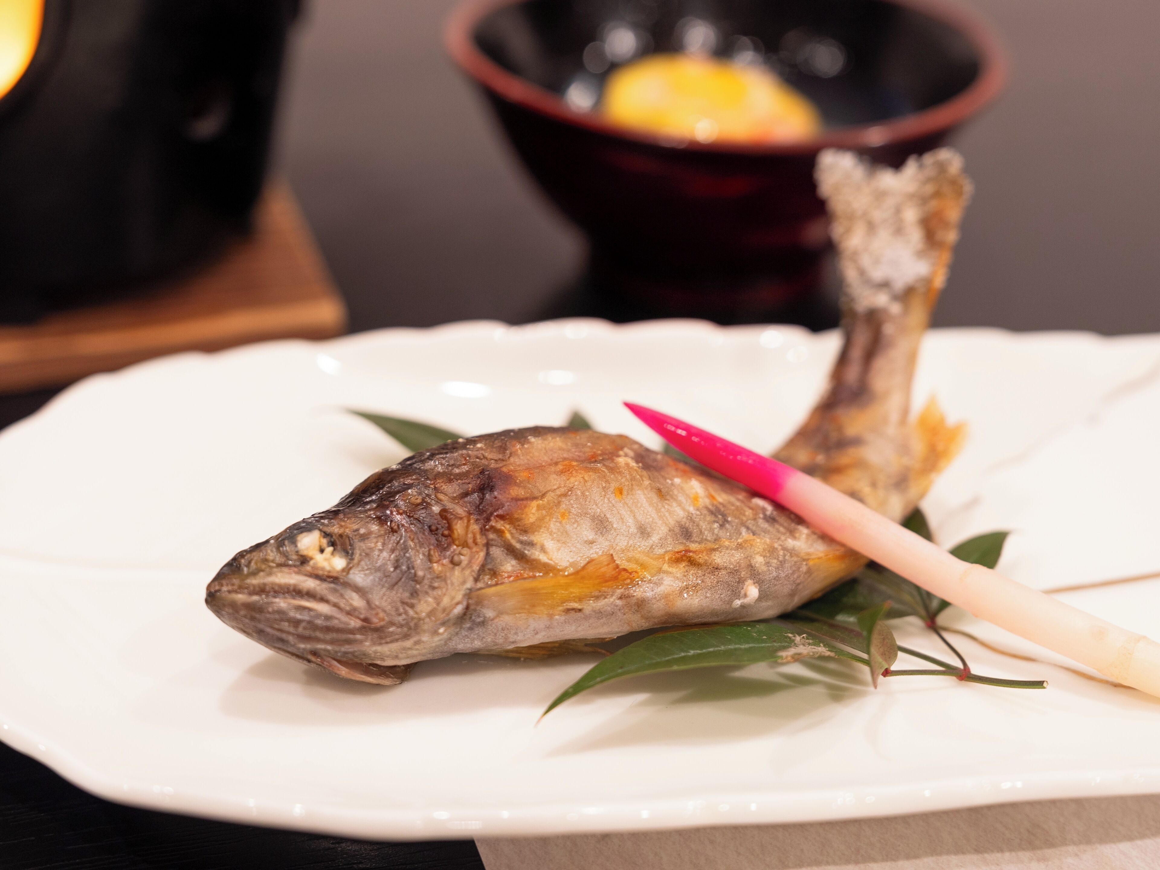 A white plate with a whole grilled fish, a pink chopstick, and green leaves.