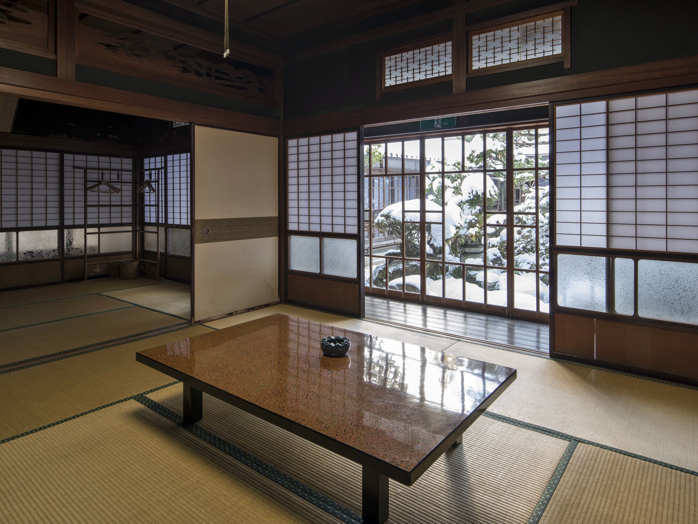 A traditional Japanese room with tatami flooring, a low table, sliding doors, and large windows with a view of a snowy garden.