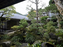 Un jardín japonés tradicional con una construcción de madera, formaciones rocosas y diversas plantas.