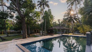 Ein Swimmingpool, umgeben von Palmen und einem kleinen Gebäude, vermutlich eine Poolhaus oder Umkleidekabine.