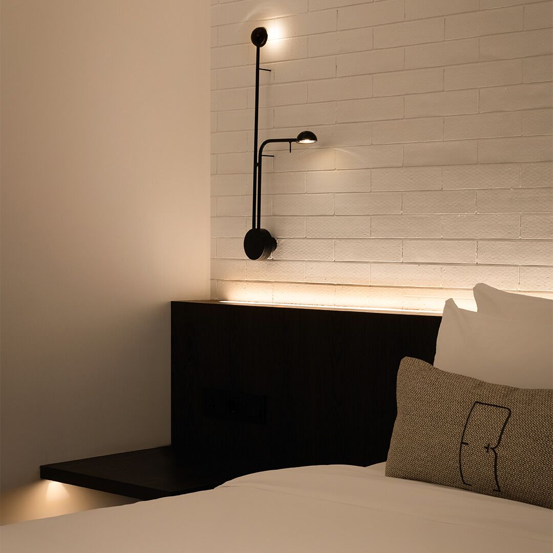 A modern bedroom with a white brick wall, a black nightstand, a bed with white bedding, and a black floor lamp.