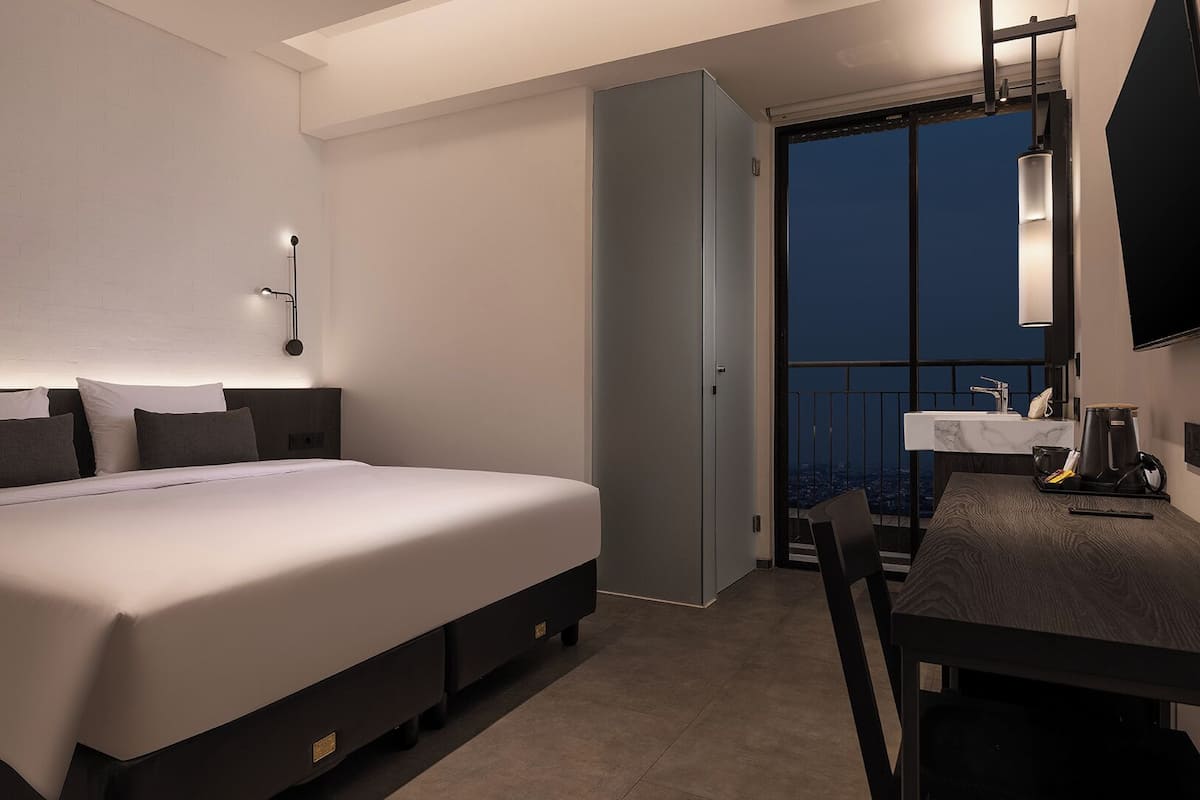 A modern hotel room with a large bed, bedside tables, a desk with a chair, a bathroom with a sink, and a view of the city at night.